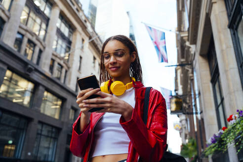 Smiling woman with eyeglasses and headphones using mobile phone in city - OIPF03560