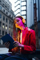 Nachdenkliche Frau hört Musik über Kopfhörer und hält einen Tablet-PC in der Abenddämmerung - OIPF03569