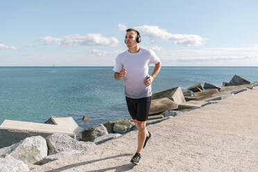 Young man wearing wireless headphones jogging on pier near sea - MMPF00980