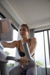 Active woman exercising on bike and using tablet PC in living room at home - IKF01395