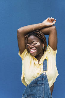Lächelnde junge Frau mit erhobenen Armen vor einer blauen Wand - PNAF06041