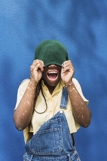 Fröhliche junge Frau, die ihr Gesicht mit einer Strickmütze bedeckt - PNAF06048