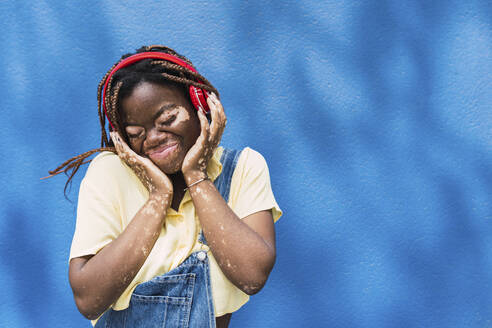 Lächelnde junge Frau mit drahtlosen Kopfhörern vor einer blauen Wand - PNAF06053
