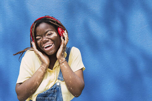 Glückliche junge Frau mit drahtlosen Kopfhörern vor einer blauen Wand - PNAF06054
