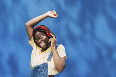 Glückliche junge Frau mit drahtlosen Kopfhörern, die vor einer blauen Wand tanzt - PNAF06055