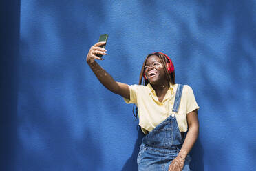 Happy young woman taking selfie through smart phone in front of blue wall - PNAF06061