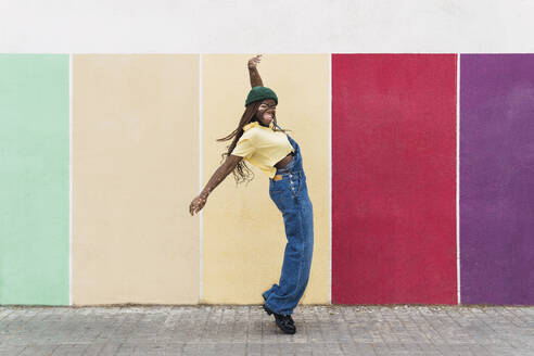 Glückliche junge Frau tanzt vor einer bunten Wand am Fußweg - PNAF06077