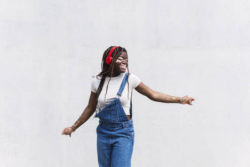 Glückliche junge Frau mit drahtlosen Kopfhörern tanzt vor einer weißen Wand - PNAF06088