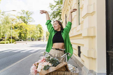 Happy woman with bicycle and arms raised on street - NDEF01208