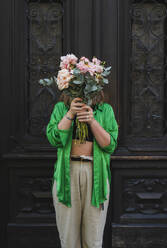 Frau, die ihr Gesicht mit einem Blumenstrauß bedeckt, steht vor einer Tür - NDEF01212