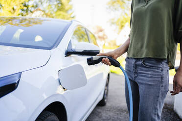 Woman holding plug near electric car at charging station - DIGF20876