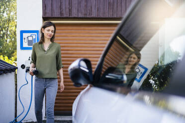 Frau mit Stromkabel neben einem Elektroauto an einer Tankstelle stehend - DIGF20881