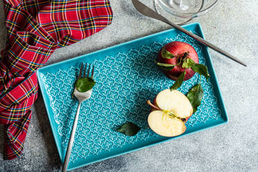 Top view of whole fresh red ripened apple with green leaves and cut half piece placed on blue tray near checkered napkin water glass on gray surface in daylight - ADSF48111