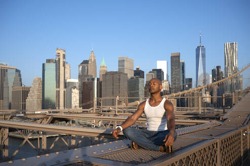 African American male sitting with legs crossed while meditating at metal suspension Brooklyn bridge infrastructure with eyes closed in daylight cityscape skyscrapers of Manhattan with blue sky - ADSF48119