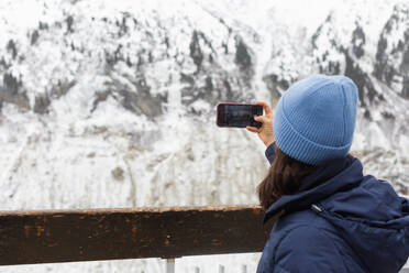 Back view of unrecognizable female standing near fence and taking photo of and snowy winter hills in Chamonix, France - ADSF48123