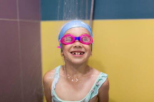 Smiling little girl swimmer with goggles and swimwear looking at camera while taking a shower after workout in pool - ADSF48124