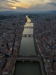Aerial view of Florence Italy in orange sundown light with bridges on Arno river and residential buildings with orange roofs against cloudy blue sky - ADSF48225