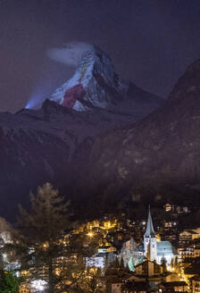 Scenic view of small settlement underneath mountain snowy peak with projector light show during night time against dark starry sky tin Matterhorn peak in Zermatt mountain resort in Switzerland - ADSF48303