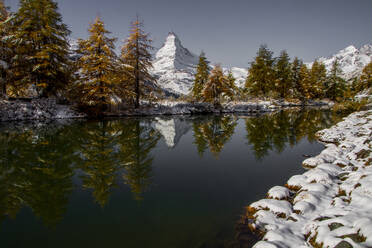 Mountains covered with snow near water lake with frozen ice surface and lush green coniferous trees on other side against blue sky on winter day - ADSF48309