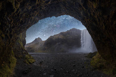 Picturesque view from cave to magnificent waterfall and rough cliff under bright sky with many sparkling stars in Skogafoss Iceland - ADSF48365