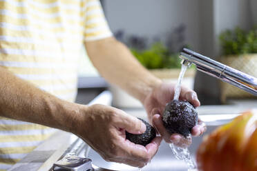 Side view of crop anonymous male washing avocado with water stream from faucet in kitchen sink while cooking in blurred background - ADSF48370