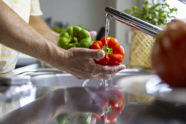 Crop anonymous male washing peppers with water stream from faucet in kitchen sink while cooking in blurred background - ADSF48372