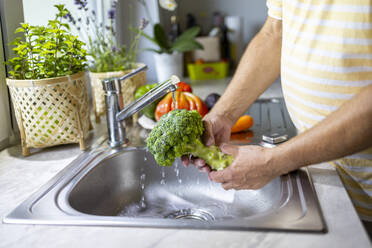 Crop anonymous male washing broccoli with water stream from faucet in kitchen sink while cooking in blurred background - ADSF48374