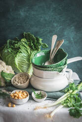 From above decorated table with various dishware ceramic bowls and spoons with fresh cauliflower, cabbage, legumes and herbs placed on table against green background - ADSF48393