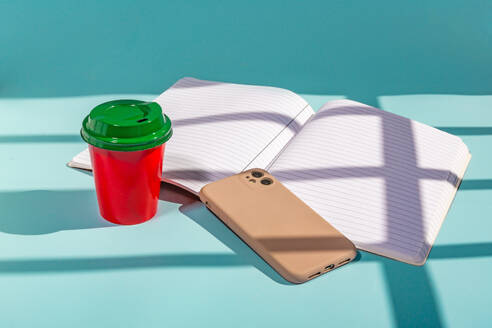 From above of minimalist red plastic cup of coffee and green lid along with a notebook and a mobile phone against blue background - ADSF48482