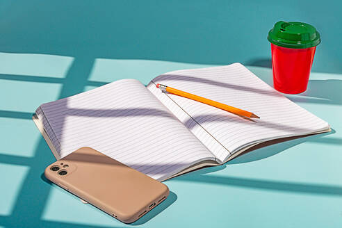 Minimalist red plastic cup of coffee and green lid along with a notebook and a mobile phone against blue background - ADSF48484