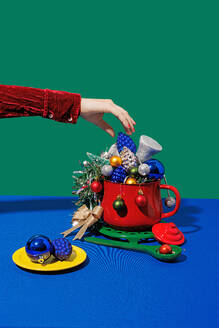 Crop anonymous female with red nails placing christmas pineapple cone in pot with different ornaments near plate and spoon against blue and green background - ADSF48487