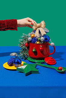 Crop anonymous female with red nails placing christmas ribbon cone in pot with different ornaments near plate and spoon against blue and green background - ADSF48488