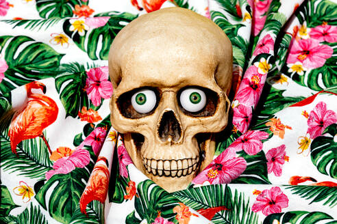 Spooky human skull looking at camera with green eyes while being placed on fabrics with drawn flowers - ADSF48493