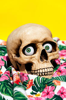 Spooky human skull looking at camera with green eyes while being placed on fabrics with drawn flowers and yellow background - ADSF48494