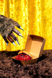 Hairy paw with metal claws of anonymous monster grabbing bloody brain with burger bun from paper box placed on fur blanket against golden curtain - ADSF48496