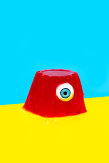 Eyeball with blue iris in red colored jelly pudding placed against vivid blue and yellow backdrop - ADSF48497