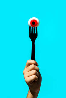 Hand of crop anonymous person holding eyeball with red iris on fork isolated on vivid blue backdrop - ADSF48498