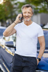 Lächelnder Mann, der über sein Smartphone spricht und sich an ein Auto lehnt - DIGF20903