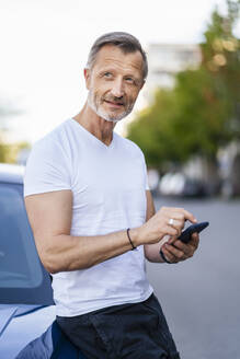 Nachdenklicher Mann mit Smartphone, der sich an ein Auto lehnt - DIGF20905