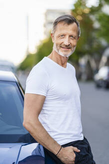 Lächelnder Mann mit Händen in den Taschen, der sich an ein Auto lehnt - DIGF20908
