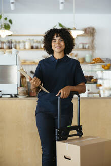 Smiling delivery woman holding clipboard and standing near package in cafe - EBSF03929