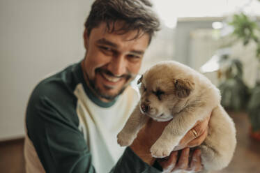 Happy man holding mixed breed puppy at home - ANAF02290