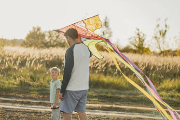 Father and son with colorful kite walking in meadow - ANAF02301