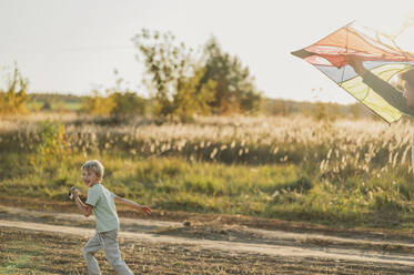 Happy boy running with flying kite in meadow - ANAF02302