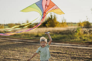 Playful boy running with multi colored kite in meadow - ANAF02303