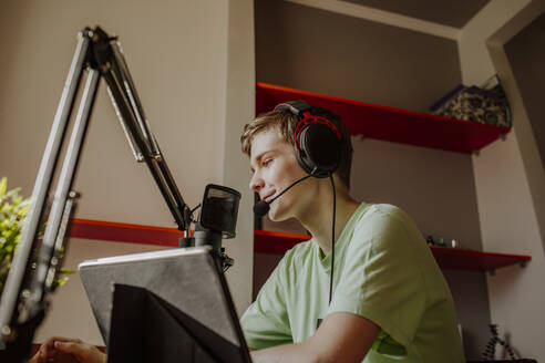 Smiling teenage boy podcasting near laptop at home - MDOF01609