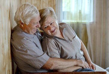 Älteres Paar umarmt sich im heimischen Wohnzimmer - MBLF00004