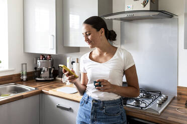 Smiling woman using smart phone and standing near kitchen at home - WPEF07724