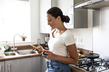 Lächelnde Frau, die in der Küche zu Hause ein Mobiltelefon benutzt - WPEF07725