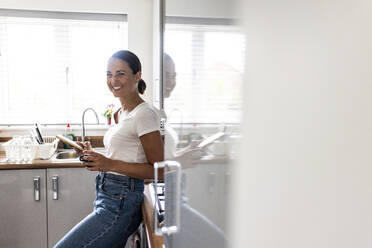 Happy woman standing with smart phone in kitchen at home - WPEF07727